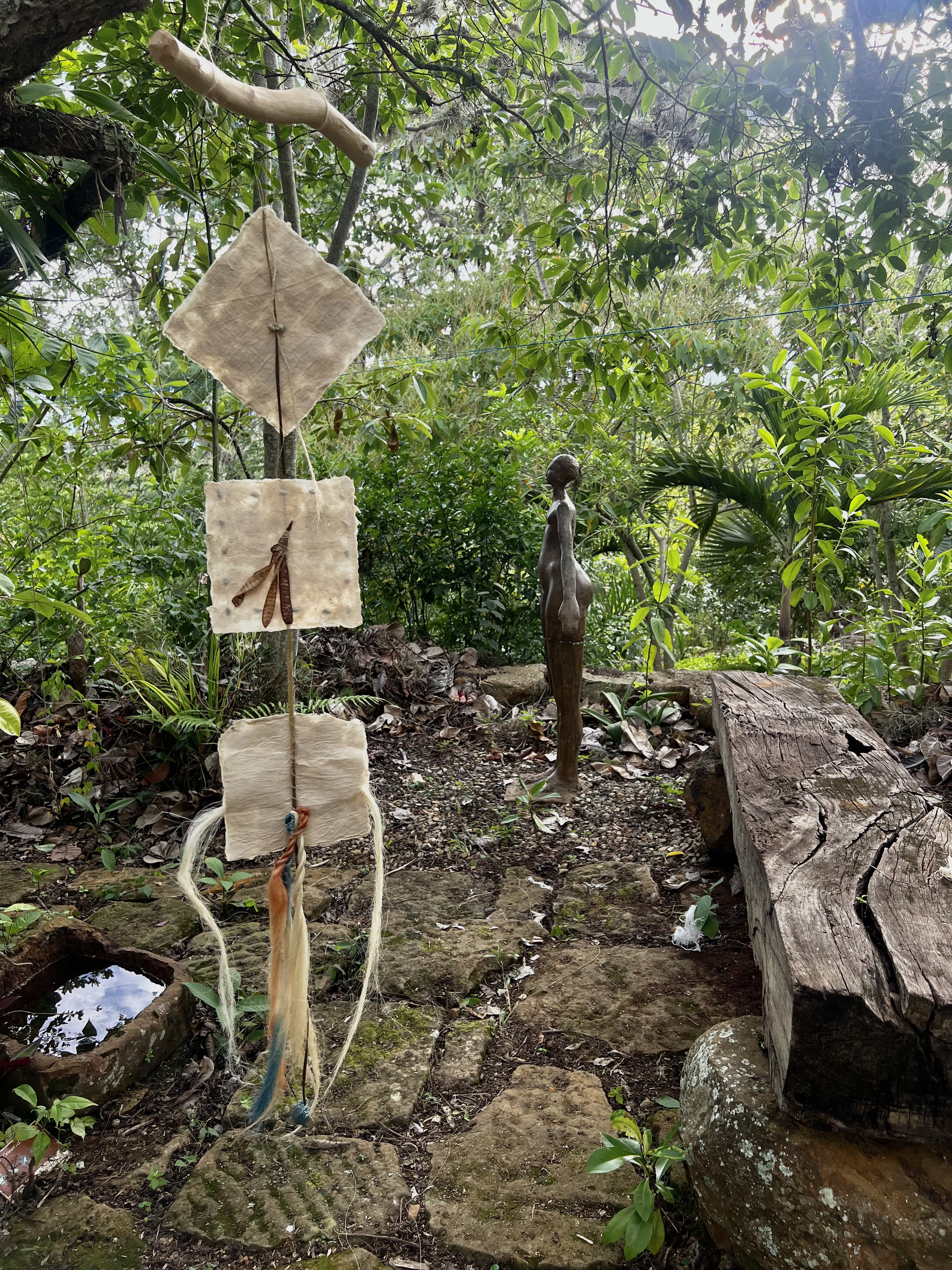 Vertical outdoor sculpture composed of square biodegradable panels and cords, placed in a forest setting.