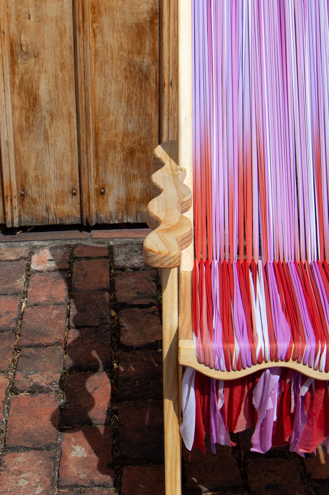 Close-up of a wooden frame sculpture with curved carved details, interwoven with vertical strands of red, pink, lilac, and white fabric strips.